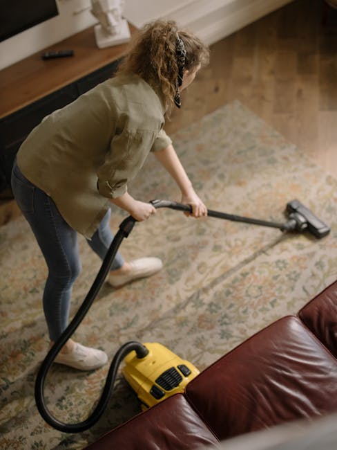 A person kneeling on a patterned carpeted floor, opening a yellow and black portable wet and dry vacuum cleaner that is connected to a flexible hose. The individual, with curly hair and wearing a tan 
