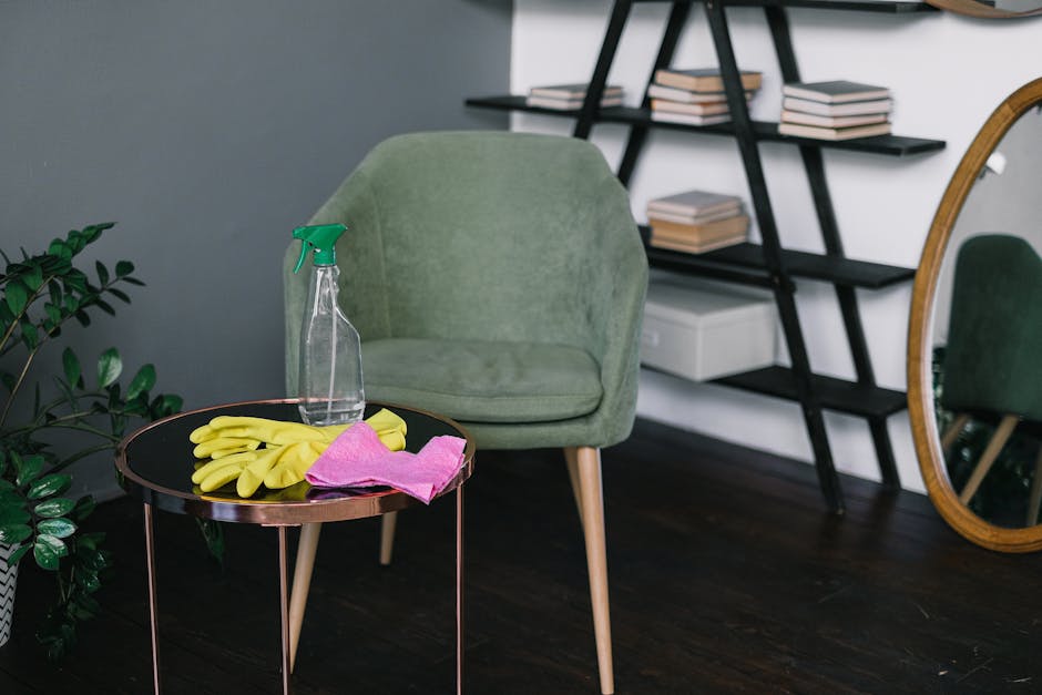 A female cleaner wearing yellow rubber gloves and a light purple top is performing surface cleaning in a domestic setting. She is reaching up to wipe or dust a hanging artificial green plant using a c