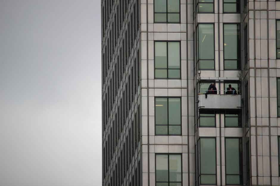 A window cleaner wearing a red jacket and safety harness is performing surface cleaning on the exterior of a high-rise apartment building in West Hampstead. The building features multiple floors with