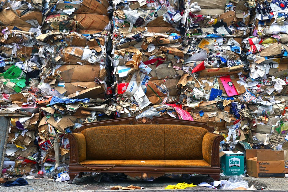 An old, vintage-style brown fabric-upholstered sofa with ornate wooden detailing on the armrests and frame, positioned in front of a large, chaotic wall of discarded cardboard and paper waste. The bac